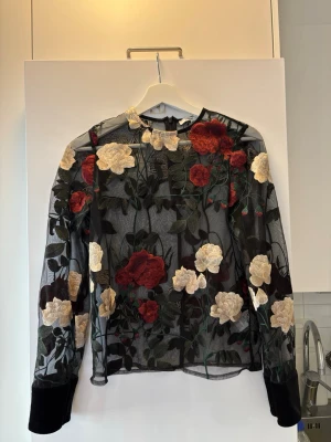 Svart meshblus med broderade blommor - Snygg svart meshblus med broderade blommor i rött, vitt och grönt. Blusen har långärmad design och transparent material som ger en trendig look. Perfekt att styla över ett linne eller topp för en cool outfit.