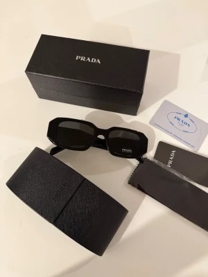 Svarta solglasögon från Prada - Snygga svarta solglasögon från Prada med rektangulära bågar och tydlig logga på sidan. Glasögonen har en chunky design och levereras med originalfodral, putsduk och certifikat. Perfekta för dig som vill ha en trendig accessoar med lyxig känsla.