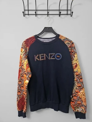 Kenzo sweatshirt med mönstrade ärmar - Cool sweatshirt från Kenzo i mörkblått med färgglada, mönstrade ärmar med tiger- och djurmotiv. Stor Kenzo Paris-logga broderad på bröstet. Tröjan har ribbade muddar och rund hals. Perfekt statement-plagg för dig som gillar unika detaljer.