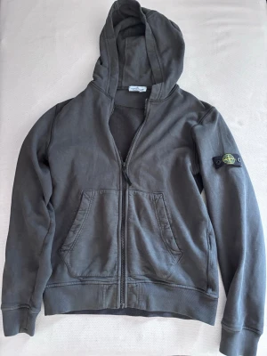 Stoneisland hoodie - Snygg mörk grå hoodie från Stone Island med dragkedja framtill och klassisk patch på vänster ärm. Hoodien har kängurufickor och ribbade muddar. Perfekt för dig som gillar streetwear och vill ha en stilren look. Tröjan är i storlek 14/170 så den passar XS - S