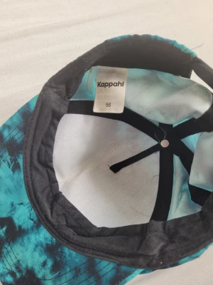 Turkos tie-dye keps från Kappahl - Snygg keps från Kappahl i turkos och svart tie-dye mönster. Klassisk böjd skärm och justerbar passform. Tillverkad i mjukt tyg som känns bekvämt på huvudet. Perfekt för att addera lite färg till din outfit.