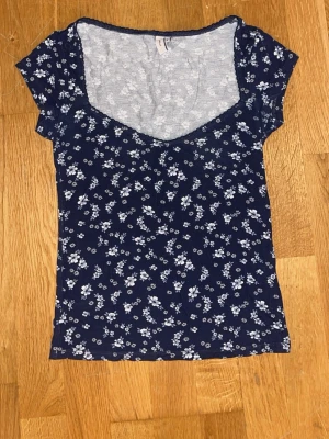 Mörkblå blommig topp med v-ringning - Söt mörkblå topp med ljusblått blommönster och v-ringad hals. Kortärmad och figurnära passform, perfekt till jeans eller kjol. Tunn och mjuk i materialet, superenkel att styla för en trendig look.