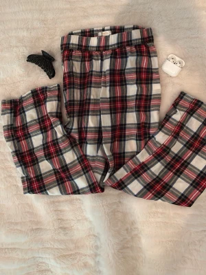 Rutiga pyjamasbyxor från Gina Tricot - Mysiga pyjamasbyxor från Gina Tricot i klassiskt rutigt mönster med färgerna röd, svart, vit och blå. Byxorna har resår i midjan och är tillverkade i mjukt bomullstyg, perfekta för chillkvällar hemma.