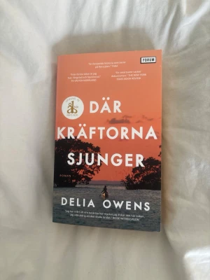 Där kräftorna sjunger pocket - Säljer romanen 'Där kräftorna sjunger' av Delia Owens. Pocketbok med omslag i orange och svart med naturmotiv och siluett av träd och vatten. Boken är utgiven av Forum och har ett prisbelönt klistermärke på framsidan.