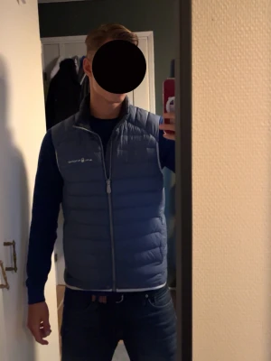 Blå dunväst från Sail Racing - Snygg blå dunväst från Sail Racing med quiltad design och dragkedja framtill. Västen har ståkrage och logga på bröstet. Perfekt att slänga över en hoodie eller tröja för extra värme. Materialet är lätt och smidigt, vilket gör den enkel att bära.