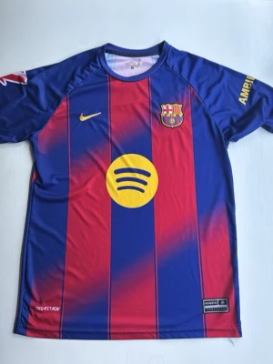 Barcelona tröja  - Snygg Barcelona matchtröja i blått och rött med gula detaljer. Tröjan har klubbmärke, Nike-logga och Spotify som sponsor framtill. På ryggen står Lamine Yamal och nummer 10 i gult. Tillverkad i lätt och ventilerande material, perfekt för fotbollsträning.