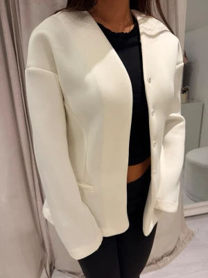 Vit stilren kavaj med knappar - Snygg vit kavaj med minimalistisk design och dolda knappar framtill. Rak passform och diskreta fickor i sidorna. Tillverkad i ett mjukt, tjockare tyg som ger en clean look. Perfekt för att lyfta vilken outfit som helst.