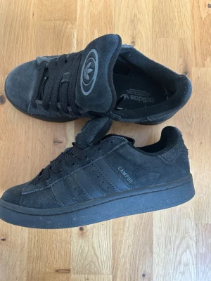 Svarta Adidas Campus sneakers - Säljer ett par svarta Adidas Campus sneakers i mocka med klassiska tre ränder på sidan och texten 'CAMPUS'. Skorna har svart sula, svarta skosnören och en stor Adidas-logga på plösen. Perfekta för dig som gillar stilrena och tidlösa sneakers. Kommer utan sin box
