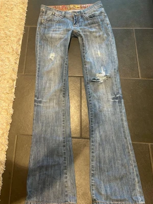 Blå bootcut jeans med slitningar - Snygga blå bootcut jeans med slitna detaljer och lätt tvättad look. Jeansen har klassisk femficksdesign och broderade bakfickor. Perfekta för en avslappnad och trendig stil. Färgen är något mörkare i verkligheten.  