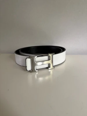 Vitt skärp med silverfärgat H-spänne - Snyggt vitt skärp i skinn med ett stort, silverfärgat H-format spänne. Bältet har en stilren och modern look med svart insida och passar perfekt till jeans eller byxor för en clean outfit.