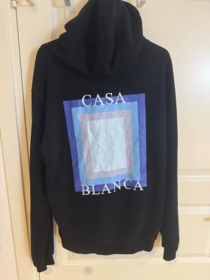 Svart hoodie med Casa Blanca-tryck - Säljer en svart hoodie med stor grafisk print i blått, lila och vitt på ryggen och mindre tryck på bröstet. Texten 'Casa Blanca' syns tydligt. Hoodien har huva och känguruficka framtill. Perfekt för dig som gillar streetwear och snygga detaljer.