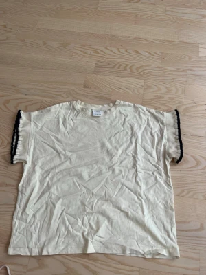 Beige topp med svarta spetsdetaljer - Trendig beige topp från Zara med korta ärmar och svarta spetsdetaljer längs ärmsluten. T-shirten har en loose passform och är tillverkad i mjukt bomullsmaterial. Perfekt för en avslappnad och stilren look.