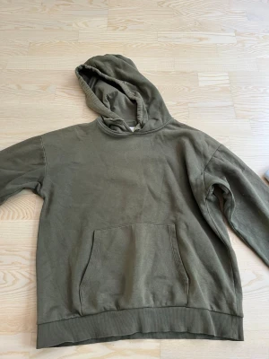 Olivgrön hoodie med magficka - En enkel olivgrön hoodie med huva och stor magficka framtill. Hoodien har ribbade muddar vid ärmslut och nederkant. Perfekt för en avslappnad och chill stil. Materialet ser ut att vara mjukt bomullstyg.
