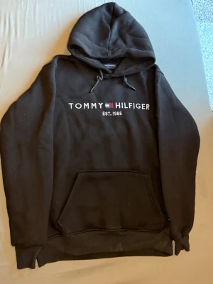 Svart hoodie Tommy Hilfiger - Säljer en svart hoodie från Tommy Hilfiger med logga och texten 'EST. 1985' broderat på bröstet. Hoodien har huva med dragsko och en stor magficka framtill. Klassisk och stilren design, perfekt till jeans eller joggers.