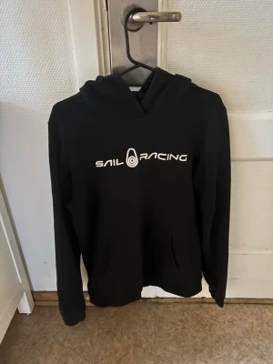 Svart hoodie från Sail Racing - Snygg svart hoodie från Sail Racing med vit logga tryckt på bröstet. Klassisk huva och känguruficka framtill. Perfekt för en avslappnad och sportig stil. Tillverkad i mjukt material som känns skönt mot huden.
