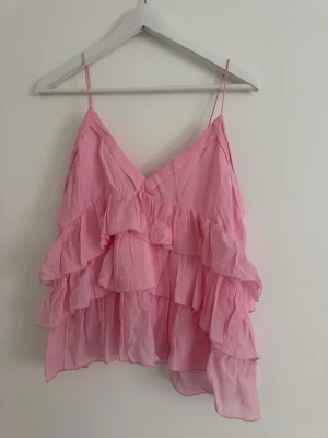 Rosa volangtop från Zara - Söt rosa topp från Zara med tunna axelband och flera lager volanger. Toppen har v-ringning och är tillverkad i ett lätt och luftigt material som passar perfekt till varma dagar. Perfekt för dig som vill ha en feminin och trendig look.