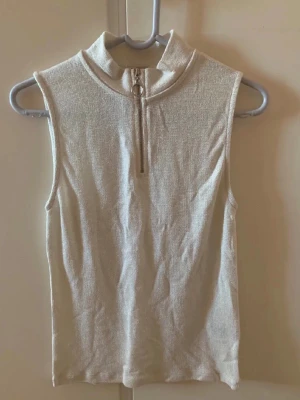 Glittrig polotopp med dragkedja - Säljer en glittrig ärmlös polotopp med halv dragkedja framtill och en snygg ringdetalj. Toppen har en lätt glittrig finish och är perfekt för dig som gillar en clean men trendig look. Passar till både jeans och kjol. Helt ny med prislapp kvar!