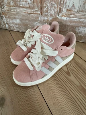 Adidas rosa Campus sneakers - Snygga Adidas Campus sneakers i rosa mocka med vita detaljer och klassiska tre ränder på sidan. Skorna har tjocka vita skosnören och en vit sula. Adidas-logga på plösen och Campus-tryck på sidan. Endast testade inomhus, tyvärr för små. Impregnerade med Axel Arigato vattenavvisande. Nypris ca 1500 kr. Storlek 39 1/3.
