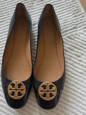 Svarta ballerinaskor Tory Burch - Stilrena svarta ballerinaskor från Tory Burch i blankt läder med rund tå och ikonisk guldfärgad logga framtill. Skorna har en klassisk och elegant design med beige innersula i läder för extra komfort. Storlek 8,5.