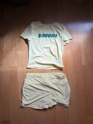Gult set med t-shirt och shorts - Säljer ett ljusgult set med t-shirt och matchande shorts. T-shirten har trycket 'SMWN' i grönt på bröstet och rund halsringning. Setet är gjort i ett mjukt och stretchigt material, perfekt för en avslappnad stil.