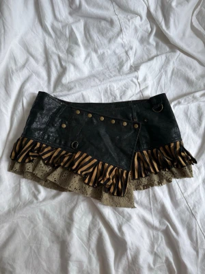 Svart minikjol med volang och nitar - Unik svart kortkjol i skinnimitation med nitar och ringdetaljer. Kjolen har volangkant i beige spets och zebramönstrad volang i svart och gult. Perfekt statement piece för dig som vill sticka ut.