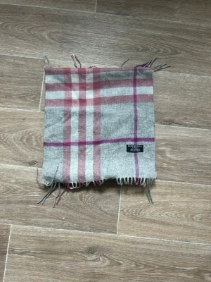 Grå-rosa rutig halsduk Burberry - Snygg halsduk från Burberry i grått med rosa och lila rutor. Klassisk rutigt mönster och fransar i kanterna. Perfekt accessoar för att lyfta din outfit och hålla dig varm. Tygmärke med Burberry-logga på ena sidan.