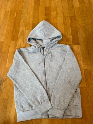 Ljusgrå zip hoodie från 157 - Ljusgrå hoodie med dragkedja från 157. Klassisk modell med huva och dragsnören, samt två fickor framtill. Finns en liten fläck som syns på bild 3