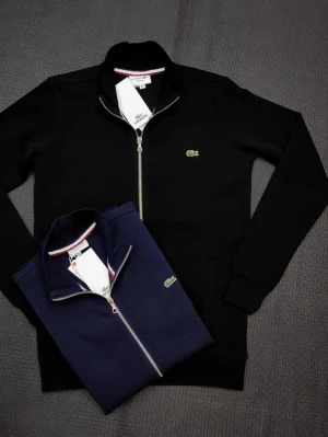 Lacoste ziptröja med logga - Snygg ziptröja från Lacoste med klassisk krokodillogga på bröstet. Finns i svart, marinblå och vit. Tröjan har hög krage, dragkedja framtill och ribbade muddar. Perfekt för en clean och sportig look. Finns både S och M storlek 
