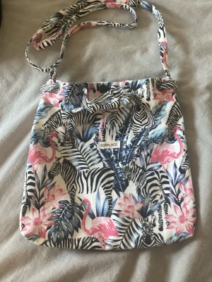Axelväska med zebror och flamingos - Axelväska i tyg med färgglatt mönster av zebror, rosa flamingos och tropiska blad. Väskan har justerbar axelrem och dragsko upptill. Perfekt för dig som gillar djurmotiv och vill ha en unik accessoar med tropisk känsla. Lite smutsig inuti men inget man ser utifrån.
