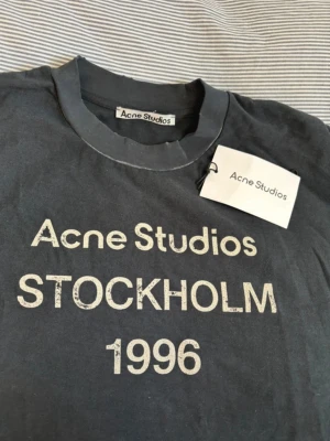 Svart Acne Studios t-shirt - Får med acne lappan .Cool svart t-shirt från Acne Studios med trycket 'Acne Studios STOCKHOLM 1996' på bröstet. Klassisk rund hals och korta ärmar. Perfekt för dig som gillar stilrena och trendiga plagg med streetkänsla.