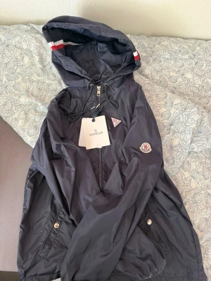 Mörkblå vindjacka från H&M - Snygg mörkblå vindjacka från H&M med huva och dragkedja framtill. Jackan har vita och röda ränder på huvan, två fickor med tryckknappar och justerbar dragsko i huvan. Tillverkad i lätt, glansigt material som står emot vind.