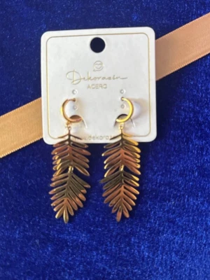 Guldiga örhängen med palmblad - Säljer ett par statement örhängen i guldfärgad metall med hängande palmbladsformade detaljer. Örhängena har en halvmåneformad ögla och ger en trendig och unik look. Perfekta för dig som vill sticka ut med accessoarerna.