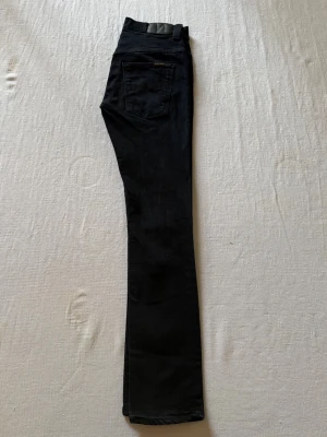 Svarta Nudie jeans - Snygga svarta jeans med klassisk femficksdesign och normal midja. Jeansen har raka ben och är tillverkade i ett stretchigt denimtyg som ger en bekväm passform. Perfekta för dig som gillar en tidlös och clean look. Storlek W29 L32!