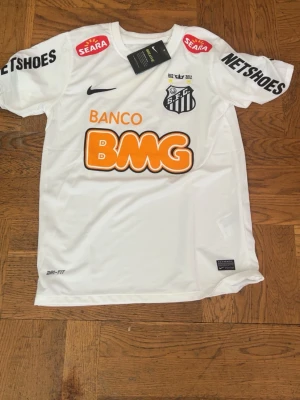 Vit Santos FC matchtröja Nike - Säljer en vit Santos FC matchtröja från Nike med klubbmärke, sponsorer och Dri-FIT-material. Tröjan har tryck i svart, orange och gult samt detaljer som jubileumsår och två stjärnor ovanför klubbmärket. Perfekt för fotbollsfans!