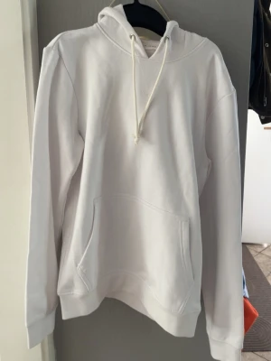Vit hoodie med huva och magficka - Klassisk vit hoodie med huva och dragsnören. Hoodien har en stor magficka framtill och ribbade muddar vid ärmslut och nederkant. Perfekt för en clean och enkel stil. Materialet ser ut att vara mjukt och bekvämt, troligen bomull eller bomullsblandning.