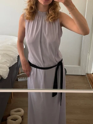 Ljuslila långklänning - Säljer en stilren ljuslila långklänning. Klänningen har en rak passform och lätt rynkad halsringning. Perfekt för dig som gillar minimalistisk och elegant stil. Svarta bältet ingår ej.