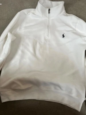 Vit half-zip tröja från Polo Ralph Lauren - Klassisk vit långärmad tröja från Polo Ralph Lauren med half-zip och hög krage. Tröjan har den ikoniska svarta polospelaren broderad på bröstet. Perfekt för en clean och stilren look.