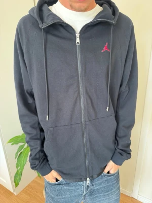 Svart zip hoodie med röd Jordan-logga - Snygg svart hoodie med dragkedja från Jordan. Har huva med dragsko, två fickor framtill och en liten röd Jumpman-logga på bröstet. Perfekt till jeans eller joggers för en sportig och avslappnad look.