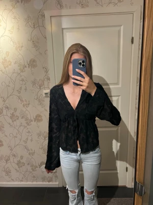 Svart blus med knyt och v-ringning - Snygg svart blus med diskret blommönster, v-ringning och knyt framtill. Blusen har långa, lite vida ärmar och lätt transparent material som ger en trendig look. Perfekt att styla med jeans eller kjol för en cool outfit. Är i storlek L men passar mig som i vanliga fall har xs/s. Aldrig använd