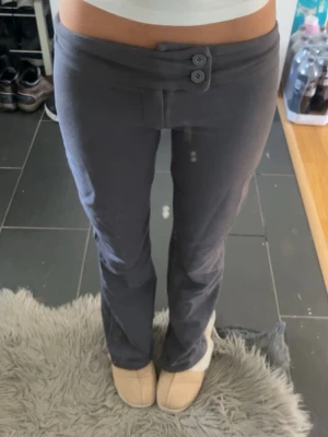 Mörkgrå kostymbyxor - Säljer ett par mörkgrå kostymbyxor från Nelly i storlek XXS. Byxorna har låg midja, bootcut och två dekorativa knappar framtill. Materialet är mjukt och stretchigt för en bekväm passform. Bild 3 ser ljusa ut men färgen ser ut som på första och andra bilden!!