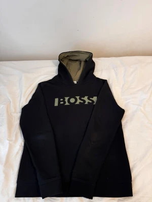 Svart hoodie från BOSS med logga - Snygg svart hoodie från BOSS med stor logga i grönt över bröstet. Hoodien har en fodrad huva i olivgrönt och ribbade muddar vid ärmslut och nederkant. Perfekt för en avslappnad och trendig look.