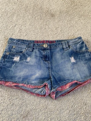 Blå jeansshorts med slitningar - Säljer ett par blå jeansshorts med slitna detaljer och rosa blommigt tyg i kanten. Shortsen har klassisk femficksdesign, bälteshällor och dragkedja. Perfekta för sommardagar och ger en avslappnad look. Shortsen är LÅGMIDJADE