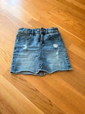 Kort jeanskjol med slitningar - Säljer en kort jeanskjol i blå denim med slitna detaljer och rå kant nertill. Kjolen har klassisk femficksdesign, knapp och dragkedja framtill samt en liten slits framtill för extra edge.