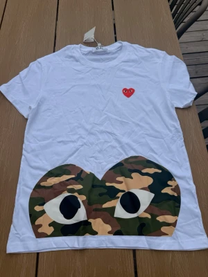 Vit t-shirt med camo-tryck och hjärtdetalj - Vit t-shirt med ett stort camo-tryck i form av ögon och hjärta på framsidan samt en liten röd hjärtdetalj med ögon på bröstet. Klassisk rund hals och korta ärmar. Perfekt för dig som gillar streetwear och unika prints.