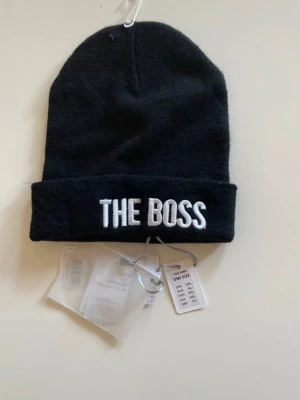 Svart mössa med texten THE BOSS - Snygg svart mössa med uppvikt kant och broderad vit text 'THE BOSS' framtill. Klassisk form som passar de flesta stilar. Tillverkad i mjukt och stretchigt material för skön passform.
