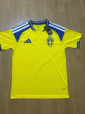 Svensk landslagströja Adidas gul/blå - Säljer en officiell svensk landslagströja från Adidas i gult med blå detaljer och vita ränder på axlarna. Tröjan har broderat Sverige-emblem på bröstet och Adidas-logga. Tillverkad i lätt och ventilerande material, perfekt för fotboll eller träning.