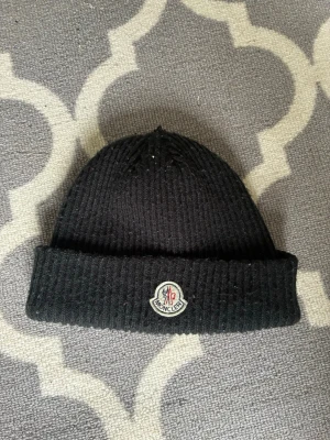Svart ribbstickad Moncler mössa - Säljer en svart ribbstickad mössa från Moncler med uppvikt kant och Moncler-logga framtill. Klassisk och enkel design som passar till det mesta. Perfekt för kalla dagar när du vill hålla stilen.