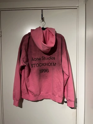 Rosa hoodie Acne Studios Stockholm - Säljer en rosa hoodie från Acne Studios med trycket 'Acne Studios STOCKHOLM 1996' på ryggen. Hoodien har en klassisk huva och avslappnad passform. Perfekt för dig som gillar streetwear och vill sticka ut med en färgstark look.