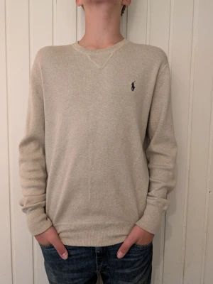 Beige tröja från Ralph Lauren - Klassisk beige tröja från Ralph Lauren med rund halsringning och diskret broderad logga på bröstet. Tröjan har ribbade muddar vid ärmslut och nederkant, perfekt för en clean och stilren look.