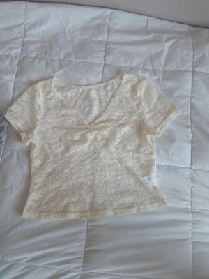 Crèmfärgad topp - Söt vit blus i spets med korta ärmar och v-ringning. Toppen har en liten rosett framtill och ett blommigt spetsmönster som ger en romantisk vibe. Perfekt till jeans eller kjol för en trendig look.